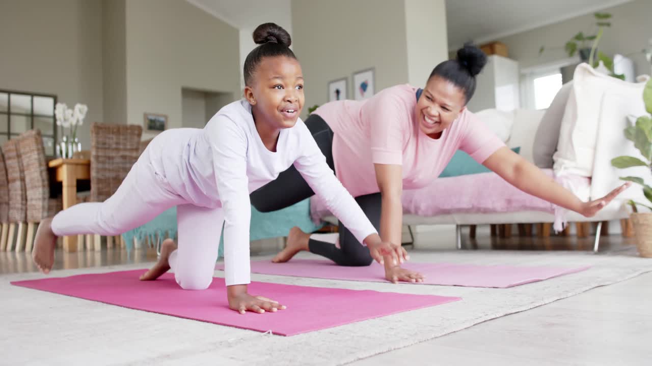 Happy unaltered african american mother and daughter doing yoga stretching at home, slow motion