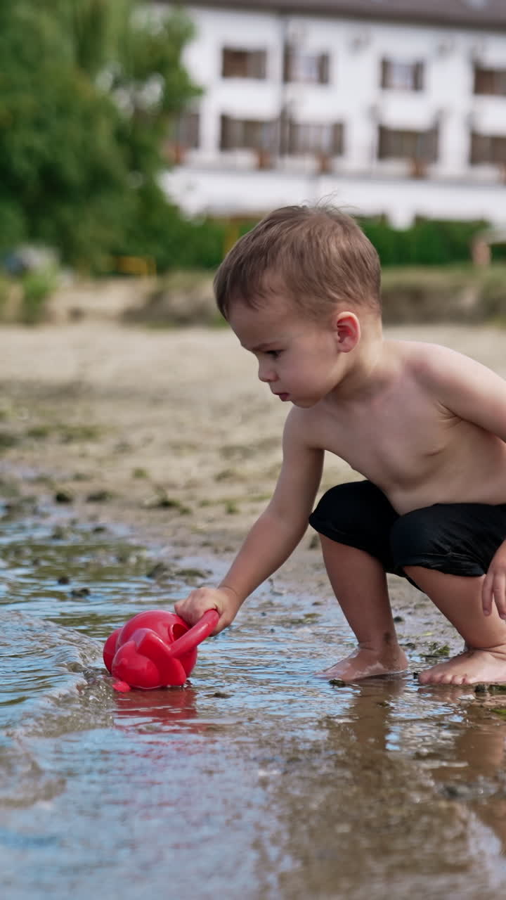 Caucasian toddler wearing black shorts filling the watering can. Baby boy playing near the river in summer. Vertical video