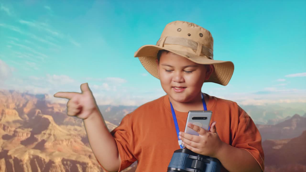 Asian Boy With A Hat And Binoculars Looking At Smartphone Then Smiling And Pointing To Side While Traveling At The Top Of Mountain. Boy Researcher Examines Something, Travel Adventure, Close Up