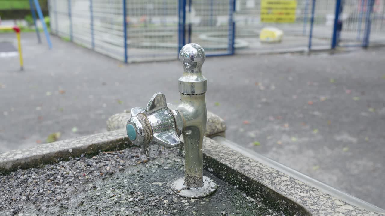 A close-up of a weathered drinking fountain in a public park, with a vintage chrome spout