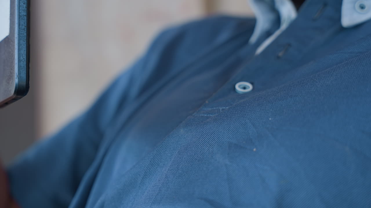 Uomo di colore con una camicia blu che scorre il telefono, primo piano della presa della mano e del tocco del colletto, sottili gesti del petto e postura che cambia sul divano, visibile la texture del tessuto e il dettaglio dei bottoni, momento di distrazione.