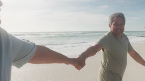 Happy-hispanic-senior-couple-holding-hands,-walking-on-beach-at-sunset