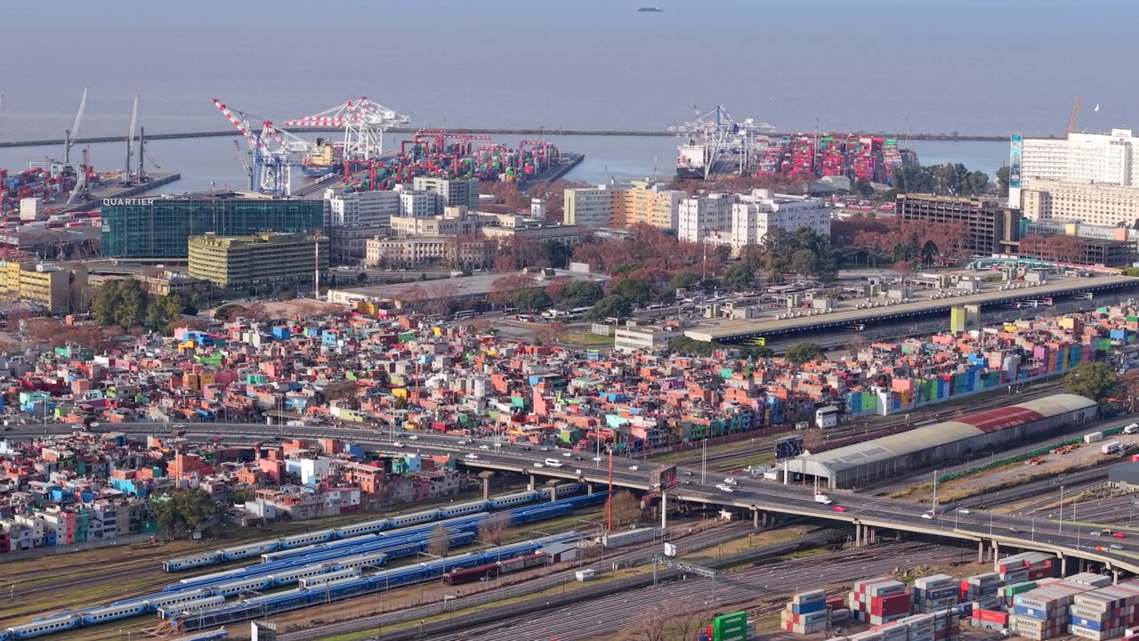 Cảng vận chuyển Buenos Aires, hiển thị các container hàng hóa xếp chồng, cần cẩu, tàu chở hàng và đường cao tốc đô thị. Cảnh quay trên không theo dõi từ trái vào một buổi chiều sáng với phông nền sông