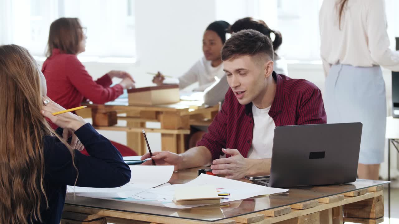 un joven feliz y sonriente hombre de negocios hablando casualmente con una colega sentada en la mesa de la oficina en un loft de coworking de moda.
