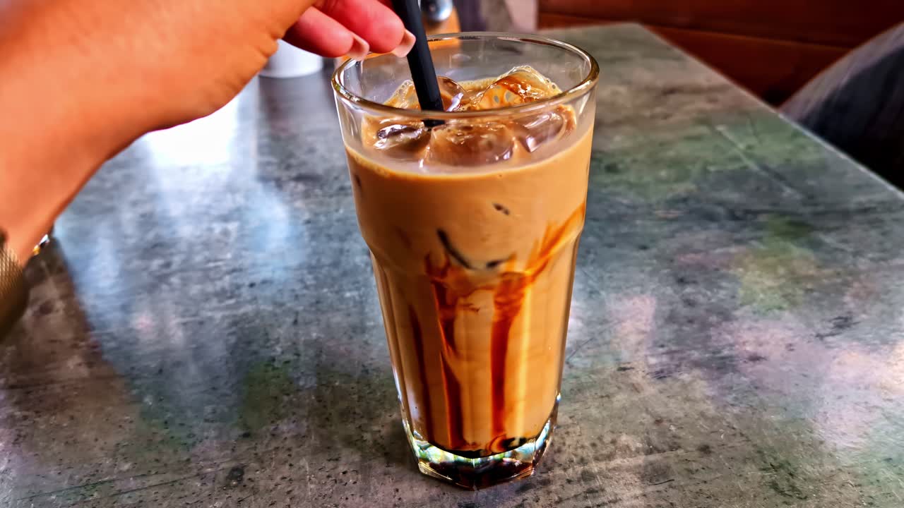 Primer plano de café caramelizado con hielo y pajita sobre una mesa texturizada de una cafetería, con una mano removiendo