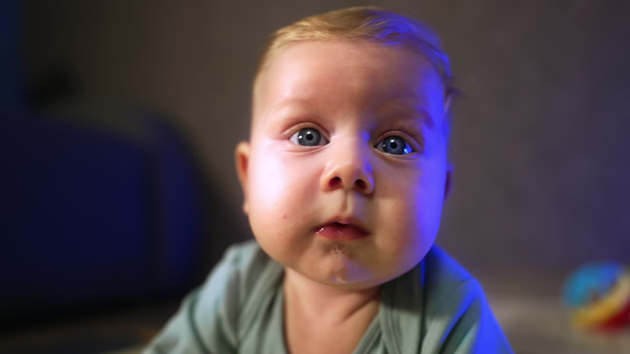 Cute face of a blonde baby with big blue eyes and plump cheeks. Close up. Infant looking ahead with surprise. Blurred backdrop.
