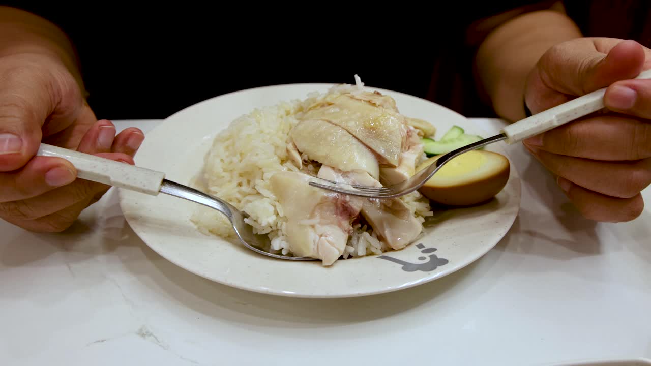 A person uses a spoon to pick up steamed chicken and rice from a white plate under bright indoor lighting, with steady camera framing