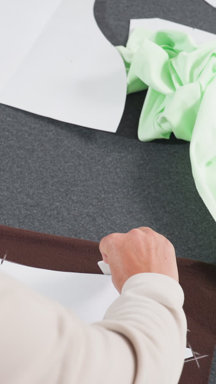 manos sujetando un patrón a un borde de tela marrón, cortador de precisión ajustando la guía de papel y alisando el margen de costura, sastre finalizando la colocación, rematador recortando el exceso cerca del mockup verde, espacio de trabajo enfocado