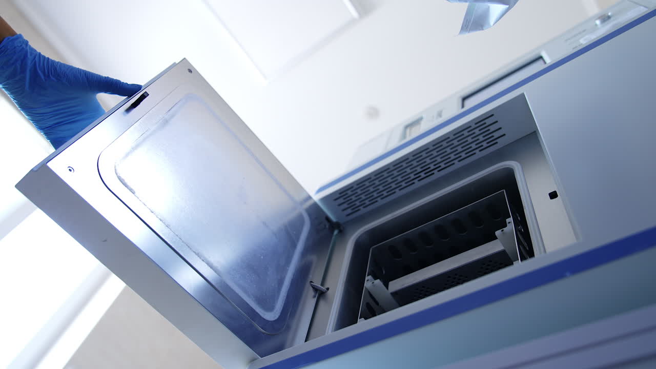 Gloved medic's hand open the door of apparatus and pulls the drawer. Nurse places the bag with tool into sterilizer machine. Low angle view.