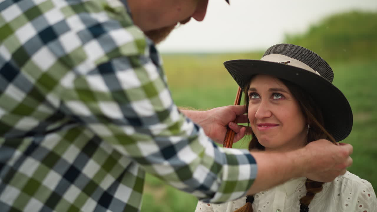 artista en una camisa a cuadros ajusta suavemente el sombrero de una mujer sonriente con el cabello trenzado, con un vestido blanco, sentado en un campo verde