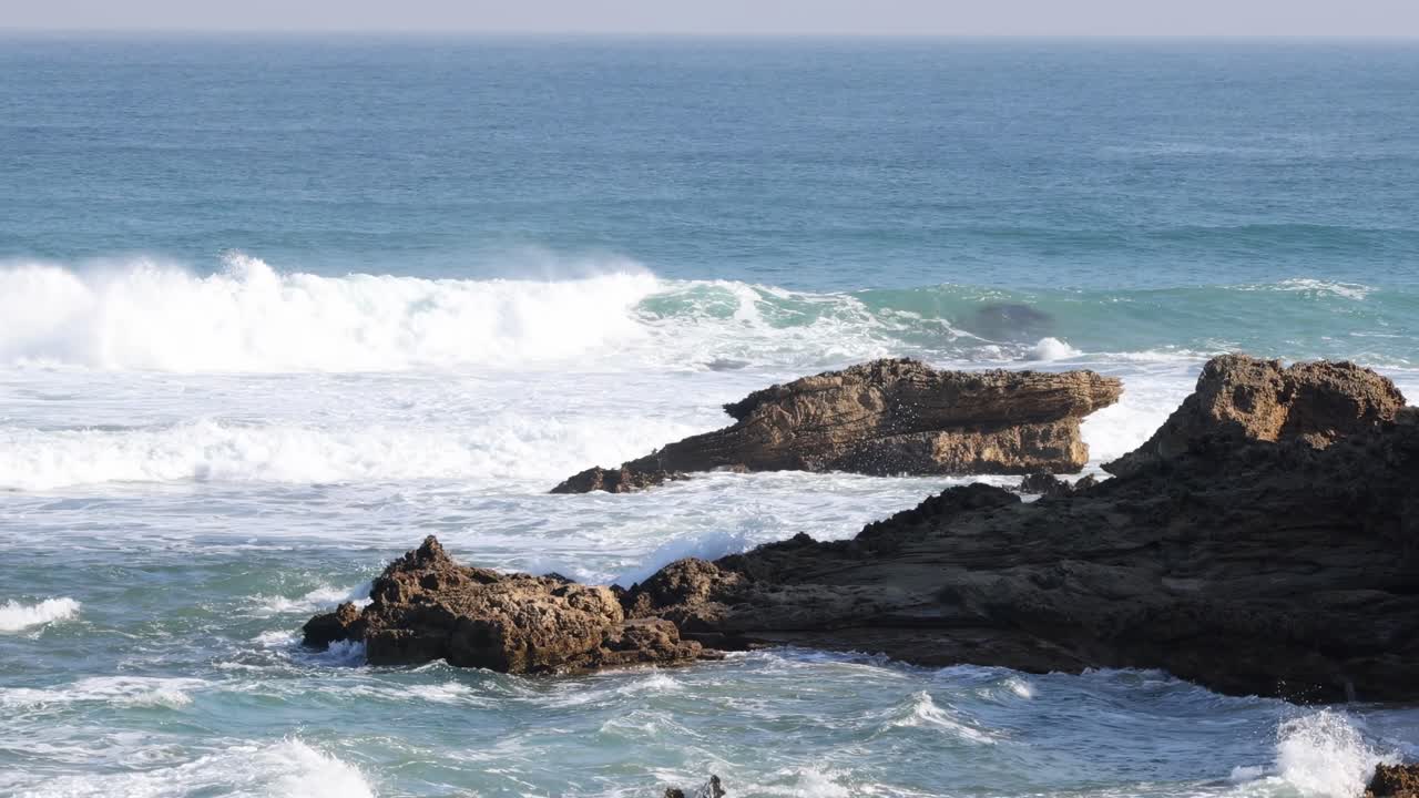 Ocean waves break against rugged rocks, creating splashes and foam under a clear blue sky.