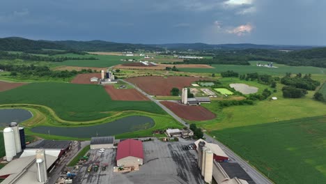 traditional american farm with silos, red barns and crop fields. surrounded by rolling hills and patchwork farmland under dramatic summer sky before storm. aerial wide shot