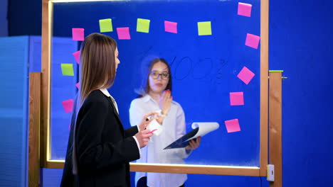 woman in black jacket points at the glass board commenting. lady in glasses holding a clipboard stands behind listening and adding few words.