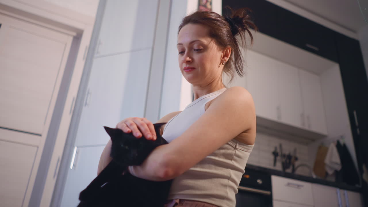 Woman Of European Descent Tends Pet In Kitchen, Contemplative Woman Prepares Meal With Her Loyal Pet Nearby, European Descent Housekeeper Gently Holds Black Cat While Cooking In Warmly Lit Kitchen
