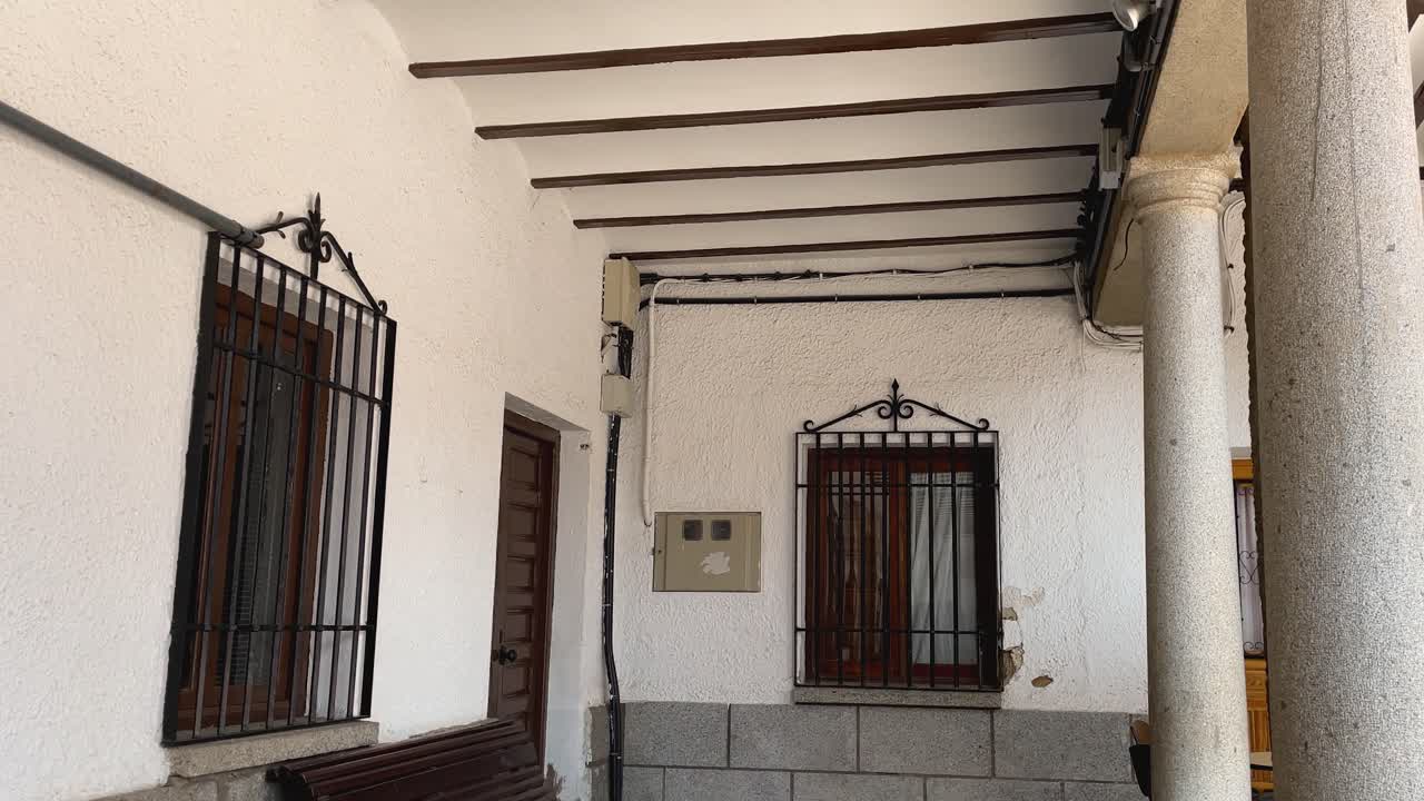 Slow-motion ascending shot of a traditional Castilian portico in Plaza Mayor, La Puebla de Montalbán, Toledo. Wooden beams, stone columns, white plaster walls, and iron grilles under autumn sunlight