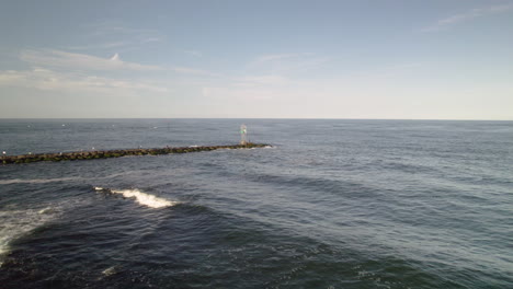 Aerial-drone-shot-pan-around-light-house-bouy-on-rock-jetty-as-man-fishes-with-heavy-lens-flare