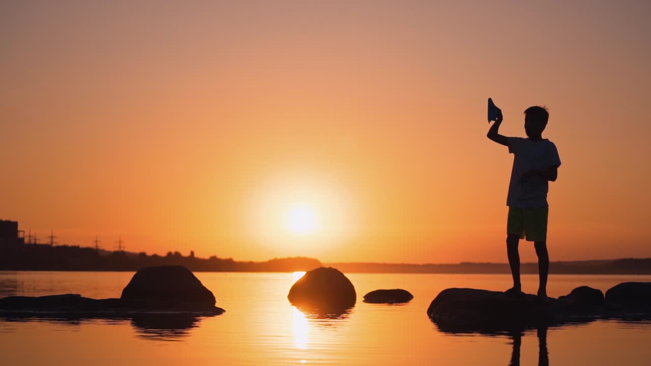 One boy playing with origami plane in summer. Silhouette of a child on a small rock among orange water at sunset. Slow motion.
