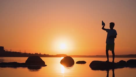 one boy playing with origami plane in summer. silhouette of a child on a small rock among orange water at sunset. slow motion.