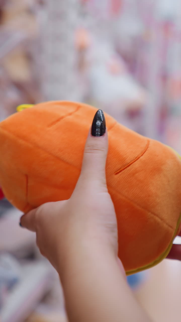 제품 세부 사항을 관찰하는 동안 상점 선반에 세련된 오렌지색 지갑을 잡는 여성의 뒷면 클로즈업, 다양한 액세서리와 프로모션 판매 표지판을 가진 소매 통로를 탐색하는 구매자