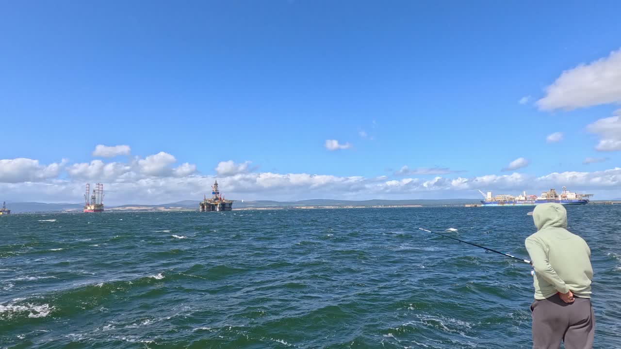 A person in a hooded jacket casts a fishing rod from a boat on the open sea near Cromarty, Scotland, under bright daylight with distant sailboats visible