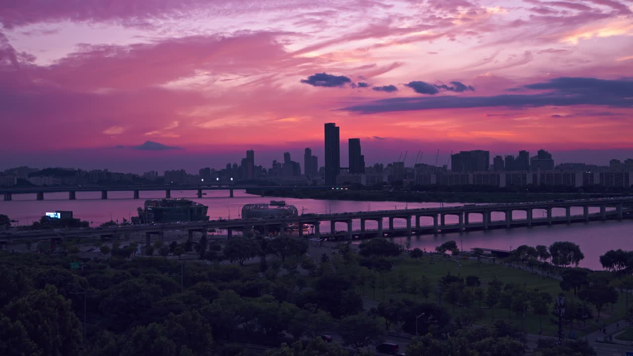 Seoul's city skyline in silhouette against a dramatic purple and pink sunset with traffic on Banpo Bridge and the green Hangang Park