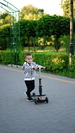 energetic toddler boy jumps cheerfully in the park. kid stands on his scooter and rides joyfully by the alley. vertical video.