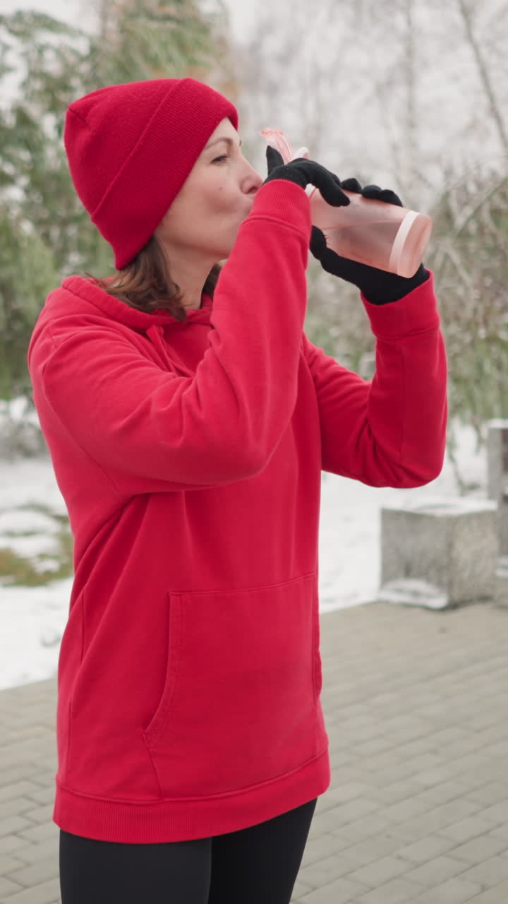 Middle-aged woman in red hoodie and beanie opens water bottle outdoors, standing in snow-dusted park with red bag on bench, surrounded by trees and serene winter atmosphere