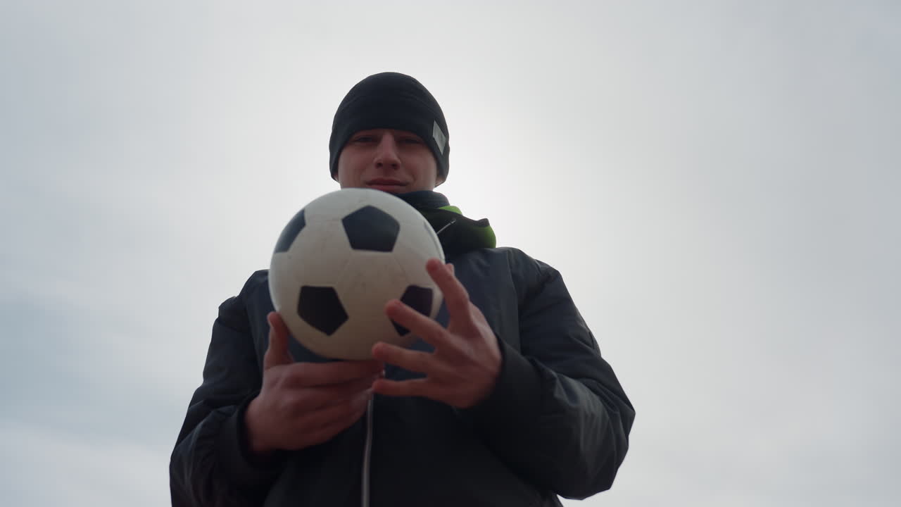 Joven atleta preparado; retrato de un joven atleta concentrado que sostiene un balón de fútbol bajo una luz solar tenue; un joven caucásico decidido agarra un balón de fútbol durante una fotografía de retrato en exteriores en ángulo bajo.