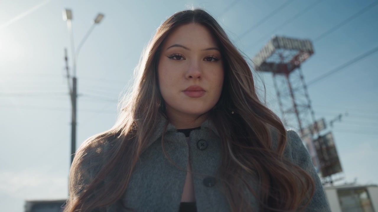 Bella artista asiatica in un ritratto all'aperto sicuro di sé con taccuino e penna, sguardo diretto, cappotto e controluce solare, torre industriale e cavi sullo sfondo, forte estetica cinematografica e posa composta.