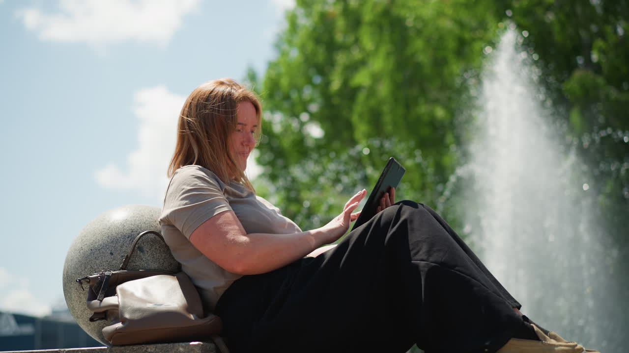 Young mum sitting outdoors by fountain admiring something on phone under bright summer sky, sunlight highlighting her hair, calm joyful mood of connection and reflection