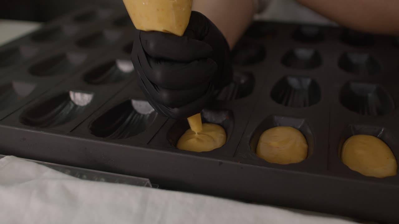 close-up shot of a pastry chef's hand in a black glove using a piping bag to fill a dark silicone mold with smooth cake batter on a marble surface