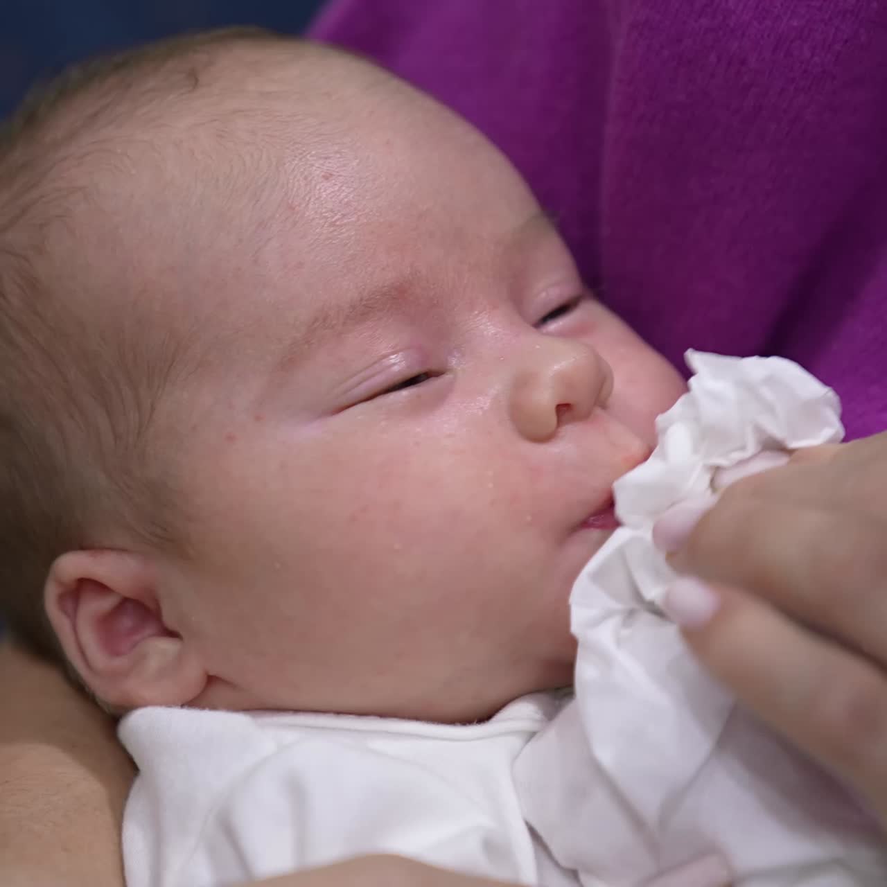Caring mother cleaning baby's mouth with a napkin. Kid grimaces as doesn't like cleaning his face. Close up