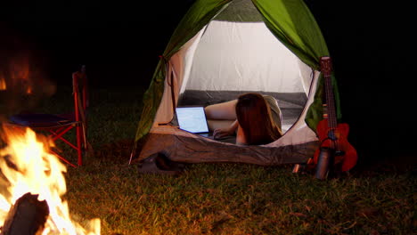 Una-Joven-Nómada-Digital-Que-Trabaja-Remotamente-Desde-Su-Tienda-Frente-A-Una-Hoguera-Por-La-Noche-Mientras-Acampa