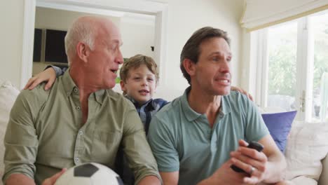 happy caucasian grandfather and adult son with grandson watching tv and supporting in living room