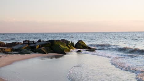 waves splash against emerald green mossy rocks and wash ashore on clear evening in slow motion