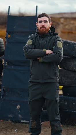 vertical video of a confident brunette man with a beard in a dark green army jacket and pants standing near tires in the steppe and posing. portrait of a confident military man standing near tires during a training exercise