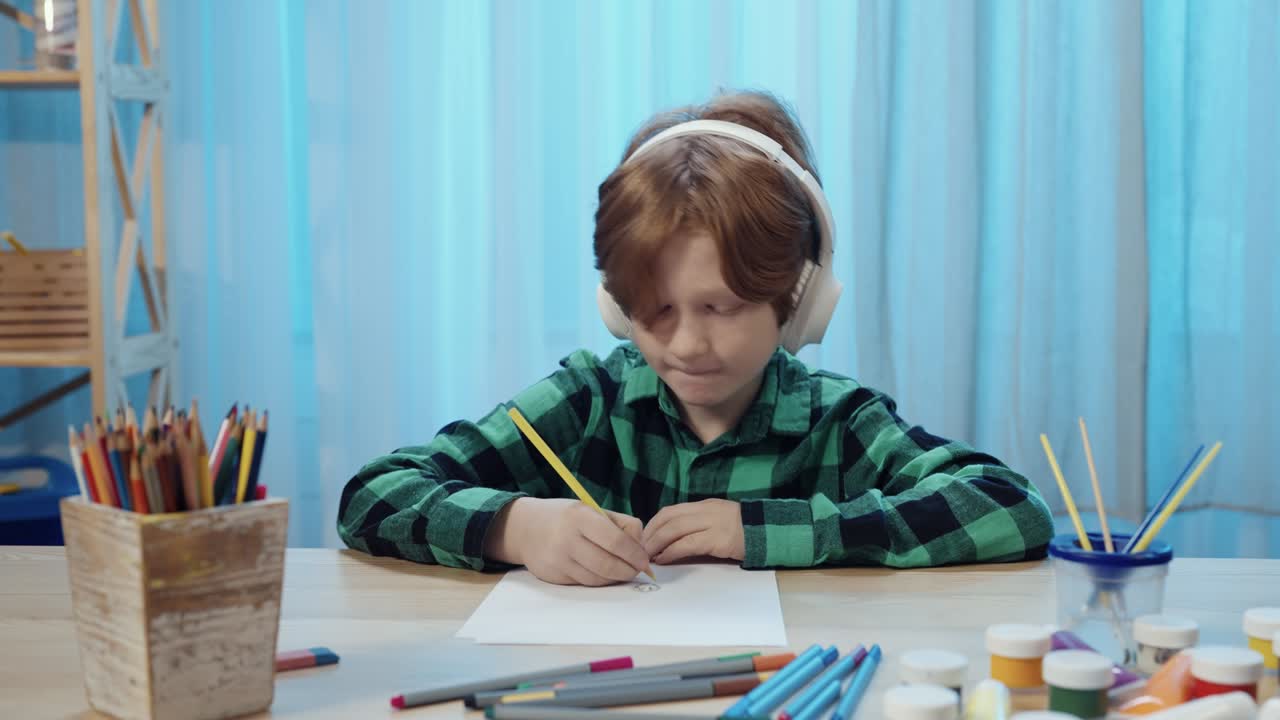 маленький школьник сидит за столом в комнате, рисует карандашом в альбоме и слушает музыку в больших белых наушниках. мальчик-подросток занимается творчеством. близко. медленное движение готово, 4k на 59,94 fps