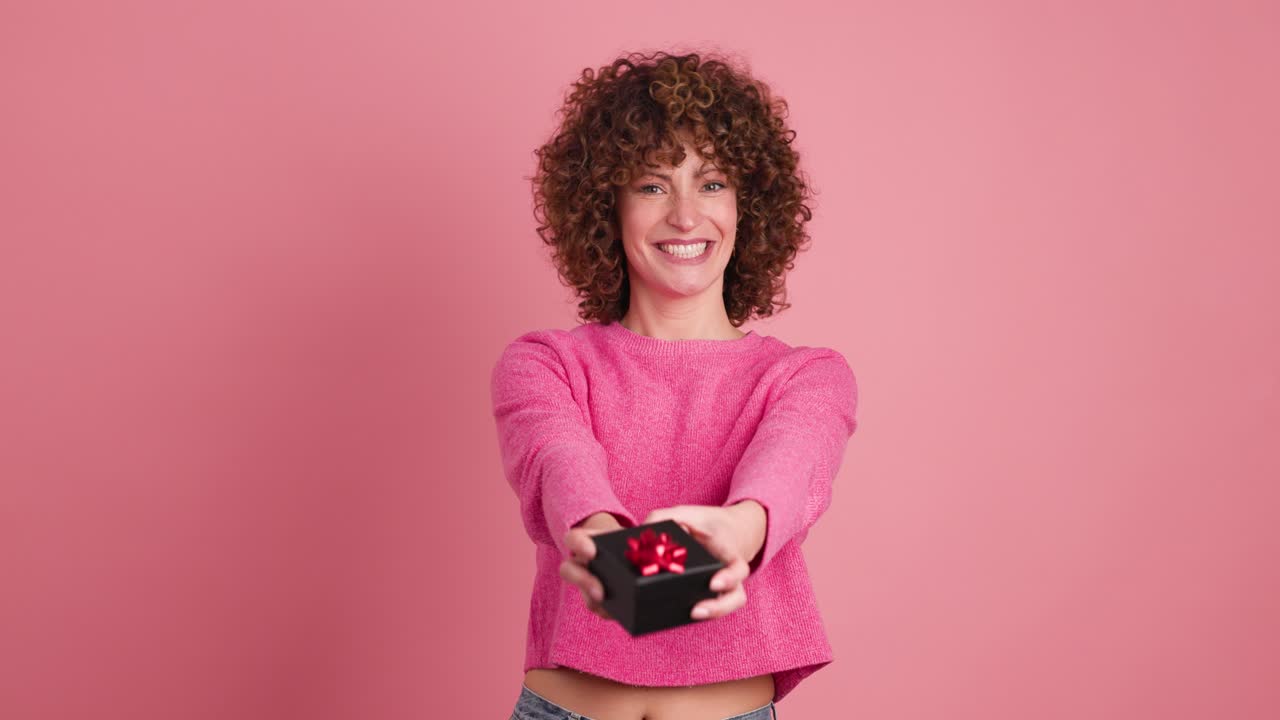 Cheerful young woman giving gift box in pink studio