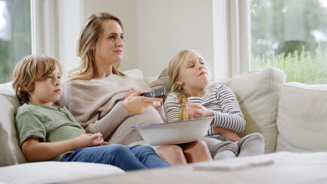 Madre-Embarazada-Con-Niños-Sentados-En-Un-Sofá-En-Casa-Riendo-Y-Viendo-Televisión-Con-Palomitas-De-Maíz