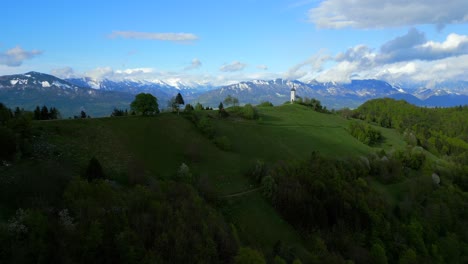 Fairytale-landscape-with-a-Jamnik-church-on-the-hill-in-the-foreground-and-a-mountain-range-in-the-background-under-a-partly-cloudy-sky