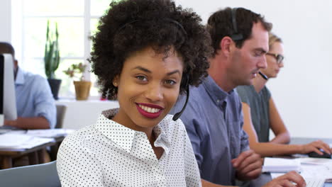 Black-woman-in-headset-smiles-to-camera-in-open-plan-office