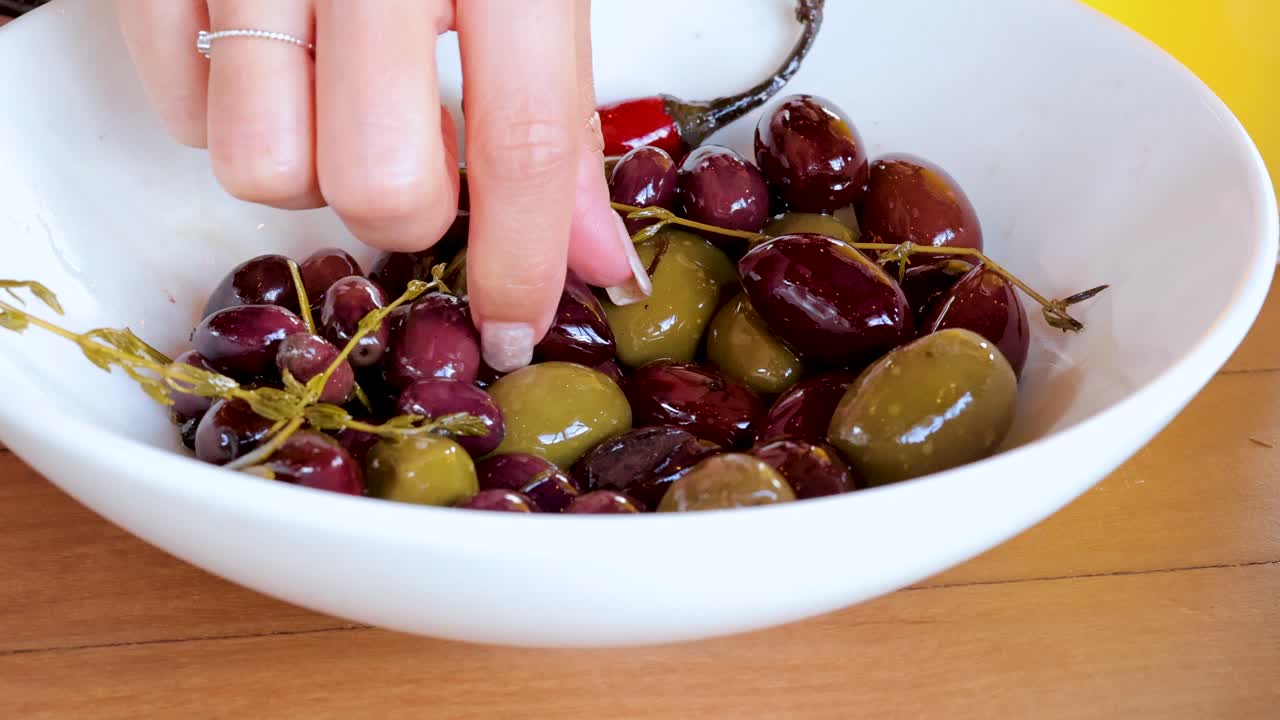 A hand and fork alternately select olives from a white bowl on a wooden table, under bright, natural lighting with a yellow background