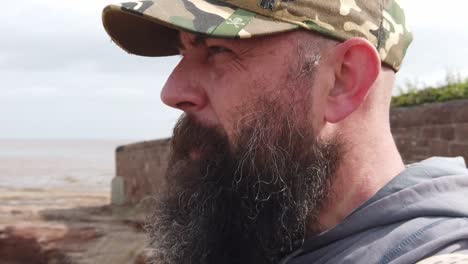 hombre urbano barbudo con gorra de béisbol mirando hacia otro lado con problemas en la playa ventosa