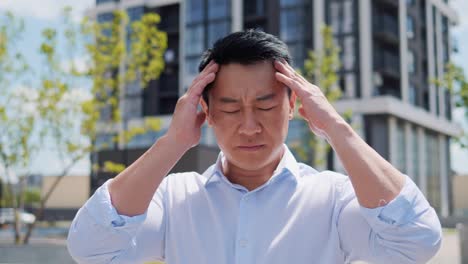 chinese tired exhausted young adult businessman, office worker having headache standing outdoors. stressed overworked male massages head. freelancer feeling terrible pain, migraine headaches.