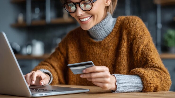 A Young Woman in a Cozy Sweater Enjoys Online Shopping with a Credit Card while Using Her Laptop at Home, Capturing the Joy of Modern Retail Therapy