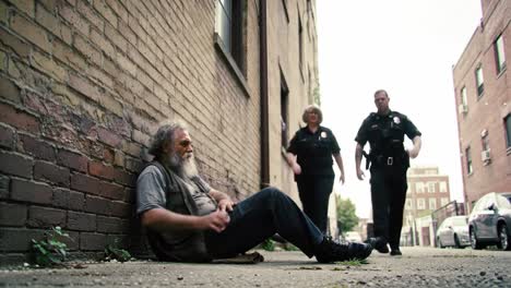 two police officers approach a homeless man sitting against a brick wall in a city alley around midday. they're engaging and ready to provide help to him.