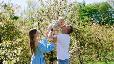 proud father holds his little son above the head. mother stands beside watching her family. beautiful parents and their child in the spring garden.