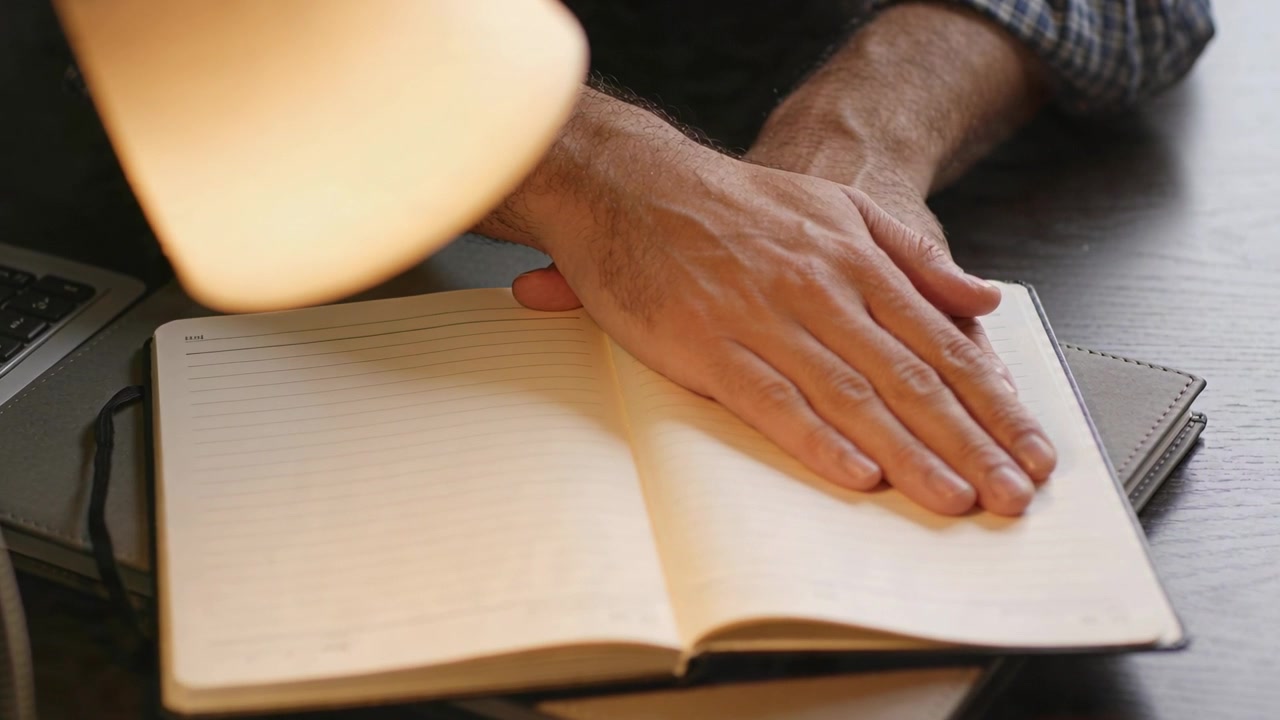 Lampe tænder, mens en højre hånd eller underarm i ternet ærme hviler på en åben notesbog og holder siderne flade.