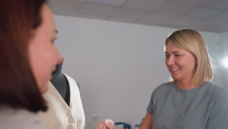 closeup shows women designers trading high five before beige vest on black mannequin, smiling brightly while celebrating fashion project success inside lively sewing studio amid fabrics and equipment