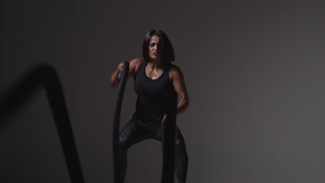 studio shot of mature woman wearing gym fitness clothing doing cardio exercise with battle ropes 3